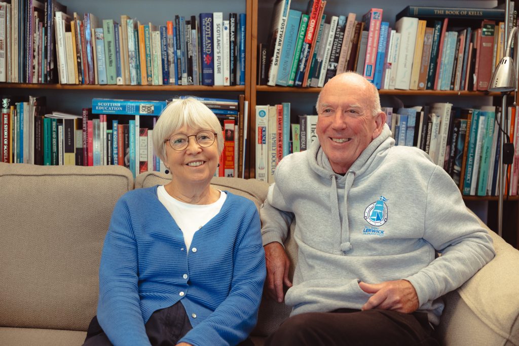 A couple sit on a sofa smiling at the camera, with bookshelves behind them.