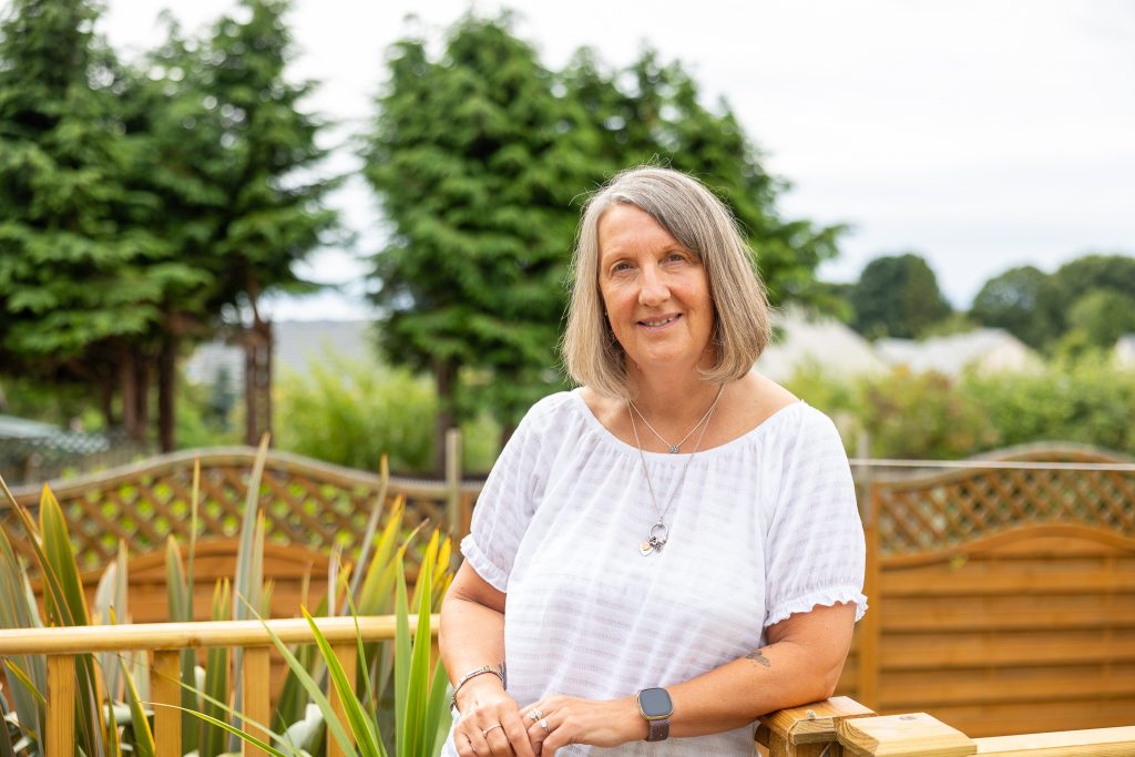 Woman standing in garden. She smiles towards the camera.